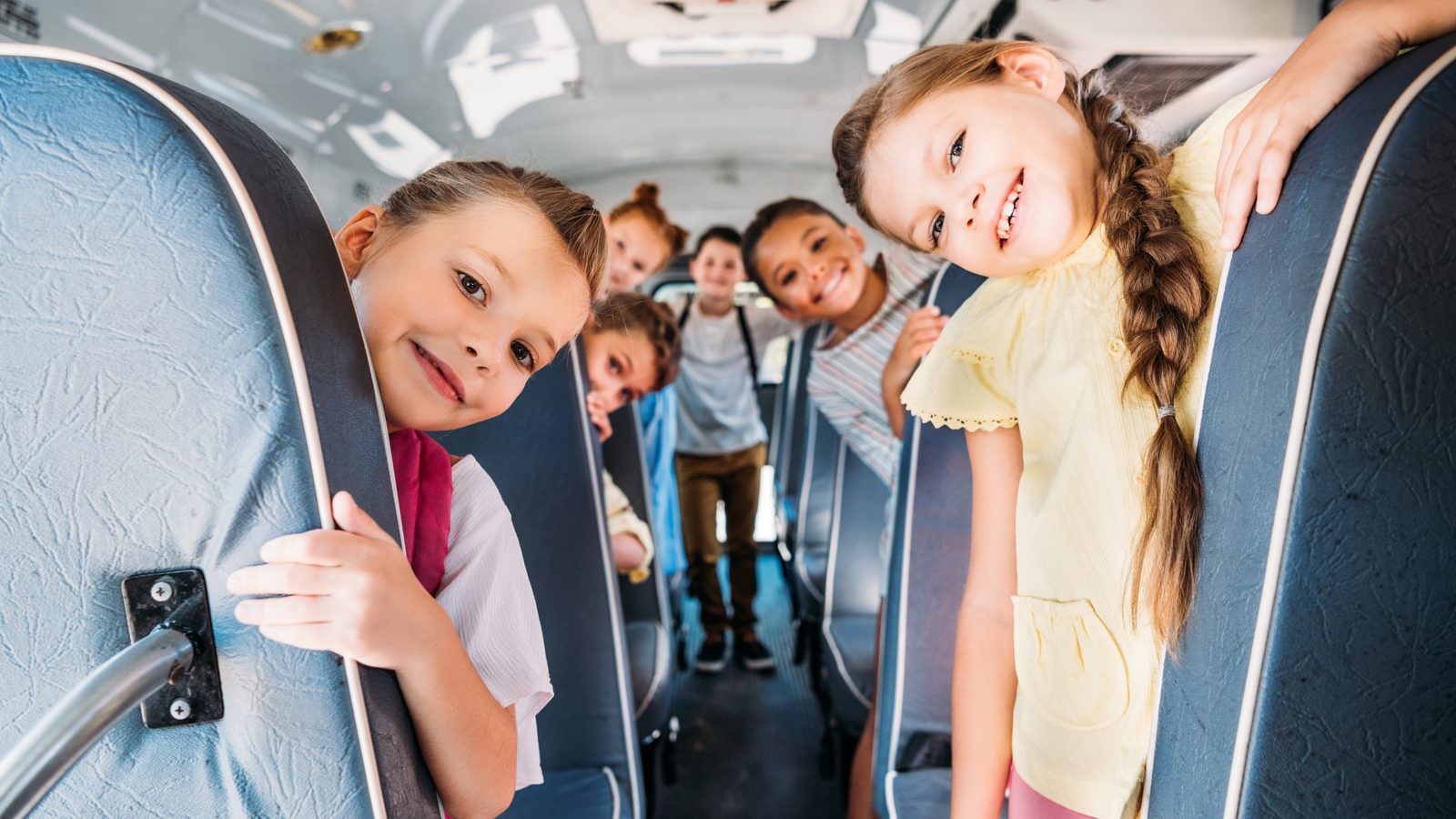 Smiling elementary students peeking out from behind the seat backs along the aisle of a school bus